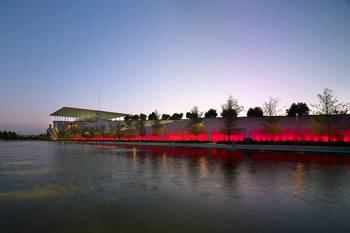 Photo showing a pond at the end of which there is an illuminated wall and a large glass building at the edge